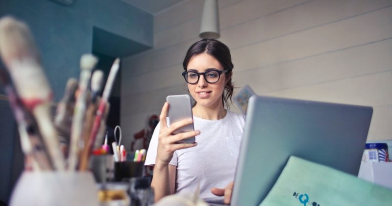 A woman in glasses is using her phone while sitting at a desk, exemplifying expectations from a logo designer.