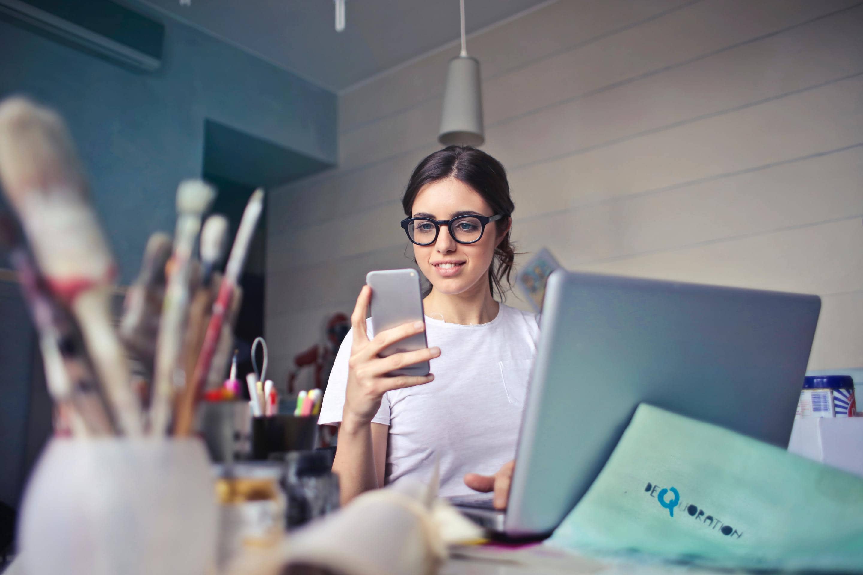 A woman in glasses is using her phone while sitting at a desk, exemplifying expectations from a logo designer.