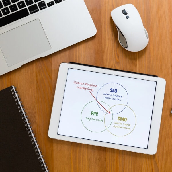 A tablet on a desk displays a Venn diagram with SEO, Pay-Per-Click (PPC), and SMO. Nearby sit a laptop, mouse, notebook, and pen.
