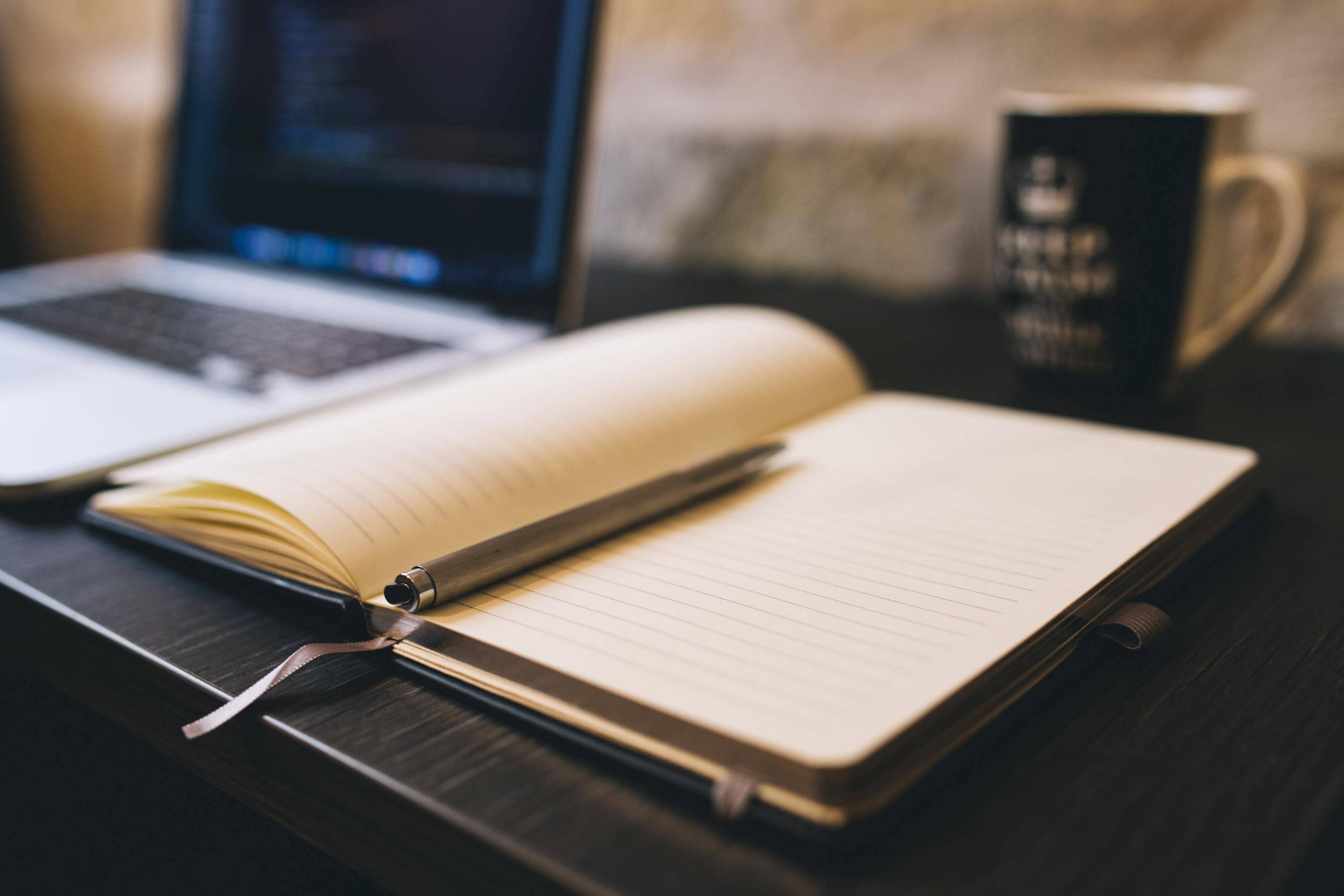 A laptop and a cup of coffee sitting beside a desk notebook.
