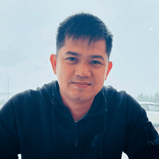 A man in a black Nike jacket sits indoors, smiling softly at the camera. The background shows a foggy window with blurred cars and trees outside.