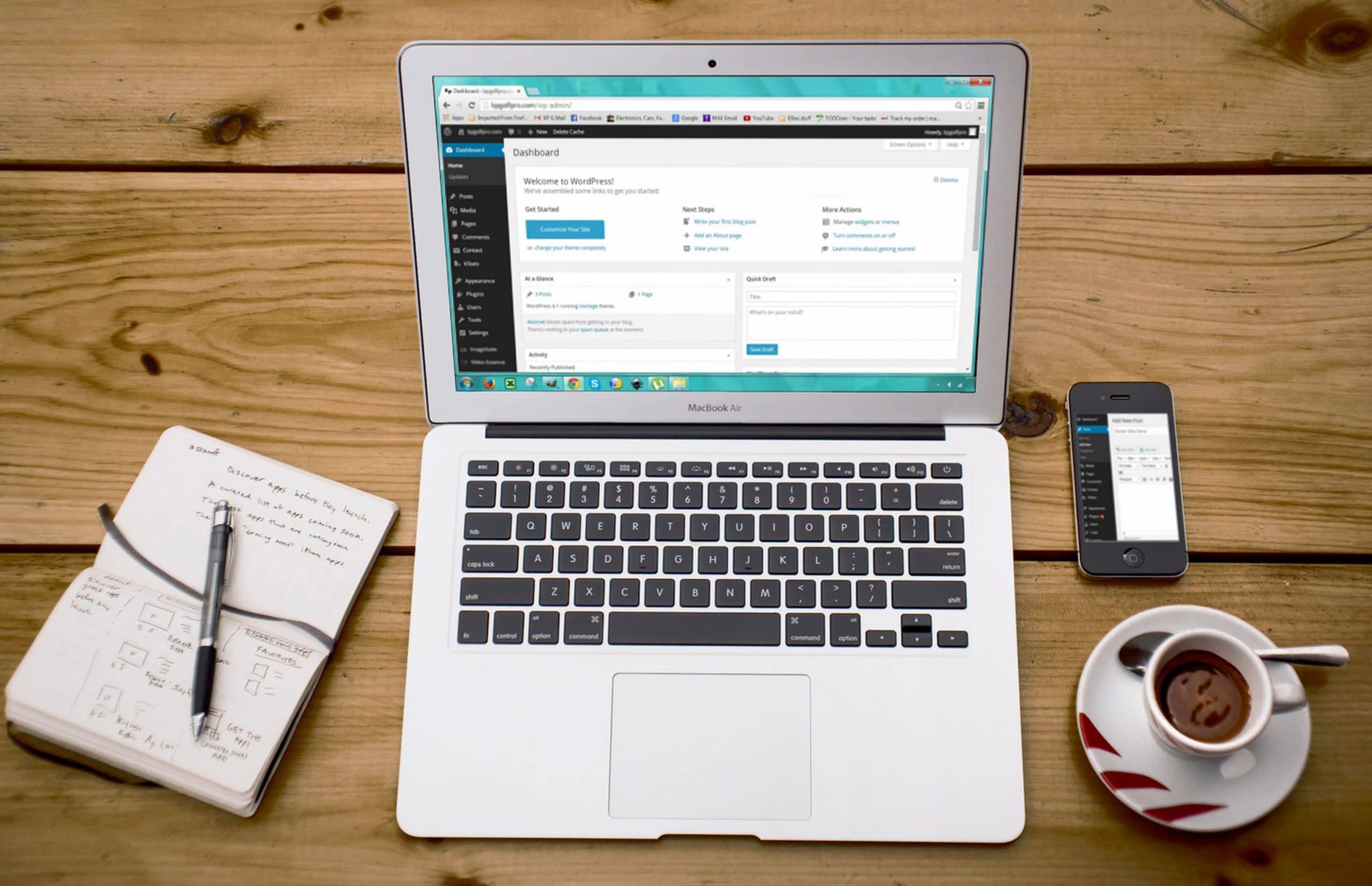 A laptop is sitting on a wooden table next to a phone and a cup of coffee.