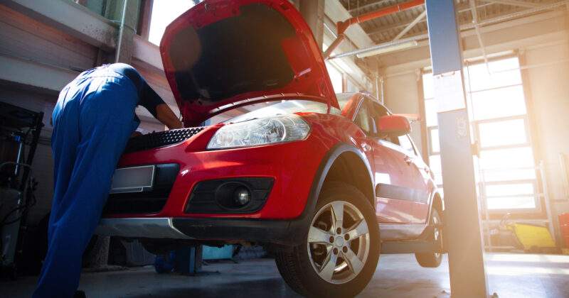 A mechanic is working on a red car in a garage while referencing the 20 Best Auto Repair Websites.