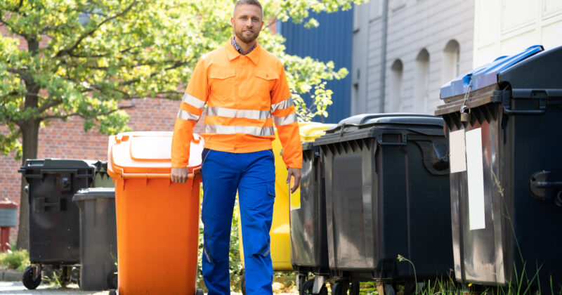 A sanitation worker in high-visibility clothing standing next to waste containers outdoors, offering Junk Removal Services.