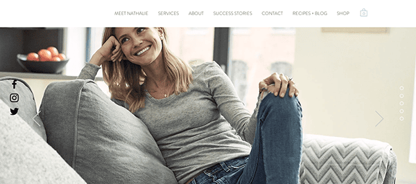 A woman smiling and sitting casually on a grey couch with her hand to her face.