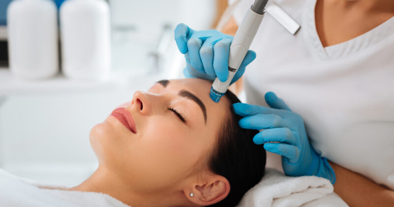 Aesthetician performing a facial treatment using a specialized device on a relaxed female client in a dermatology spa setting.