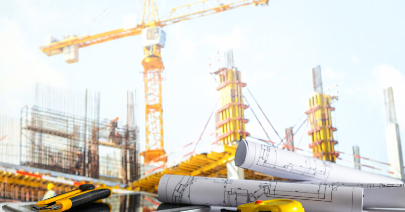 Blueprints and civil engineering tools in front of a busy building site with cranes and scaffolding.