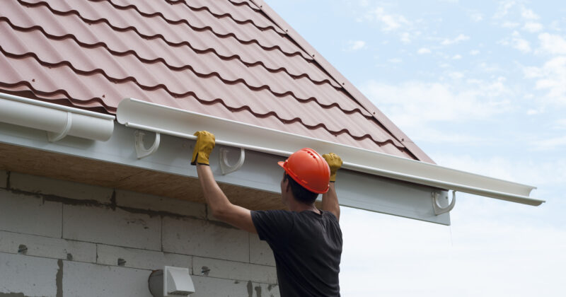 A construction worker installs a white rain gutter on a house with a red tile roof. The worker, donning a red hard hat and yellow gloves, is following the latest trends in gutter website designs to ensure optimal functionality and aesthetic appeal.