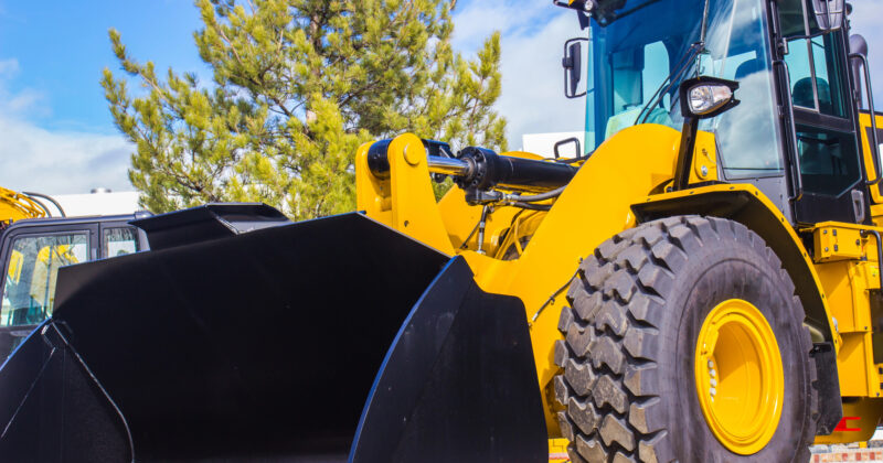 Yellow heavy equipment wheel loader with a large bucket parked outdoors, with trees in the background under a blue sky.