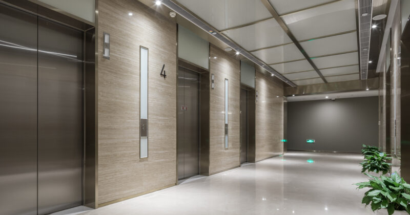 A modern, well-lit elevator lobby with four elevators, indicated by the numbered signs above each door. The area, reminiscent of a professional therapy practice, is decorated with potted plants and has a polished marble floor.