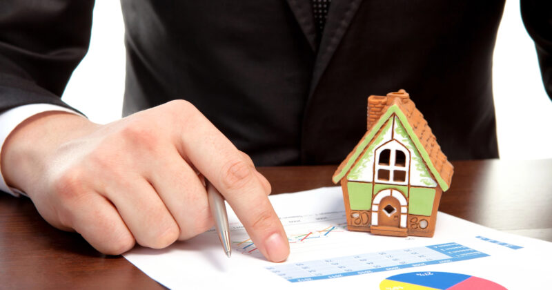 A person in a business suit signing documents with a small model house next to them, symbolizing a real estate transaction on one of the best property management websites.