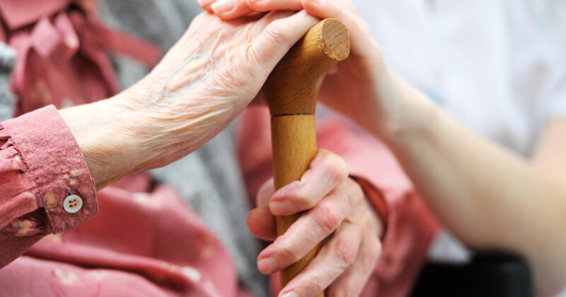 An elderly woman's hands clasp a wooden cane, comforted by a younger person's hands on top, depicting a caring gesture in a senior living design.