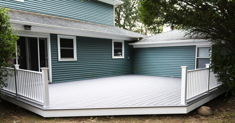 Spacious white deck designed by the best deck builders, attached to a blue house with multiple windows and a sloping roof, surrounded by greenery.