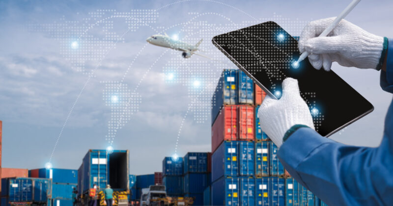 A person wearing gloves uses a tablet to manage logistics through a website design, with an airplane and digital map overlay, in a shipping container yard.