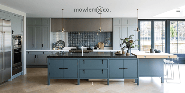 A modern kitchen with blue cabinetry, a large central island, and stainless steel appliances. The space features a mix of open and closed storage, with large windows providing natural light.