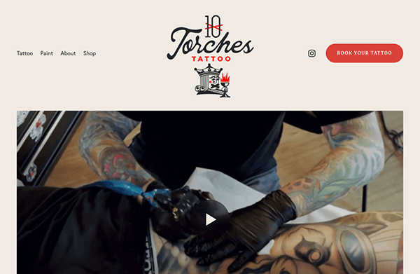 Tattoo artist working on a client's arm, seen from above, in a tattoo studio with colorful ink and equipment visible.