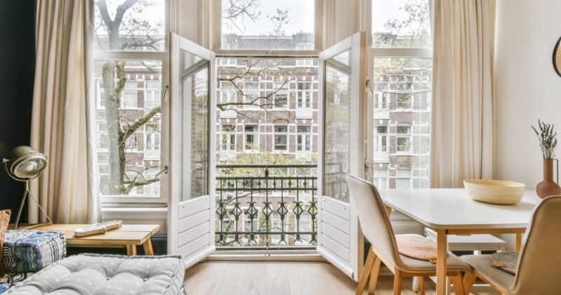A bright living room with open French doors leading to a small balcony. The room features a cushioned sofa, a table with chairs, and unique window designs offering a view of the neighboring building through the windows.