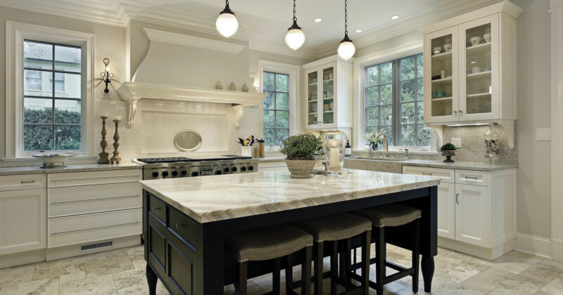 A modern kitchen with the best kitchen cabinets, a large island featuring a marble countertop and stools, stainless steel appliances, and three pendant lights. The room has large windows and various decorative accents.