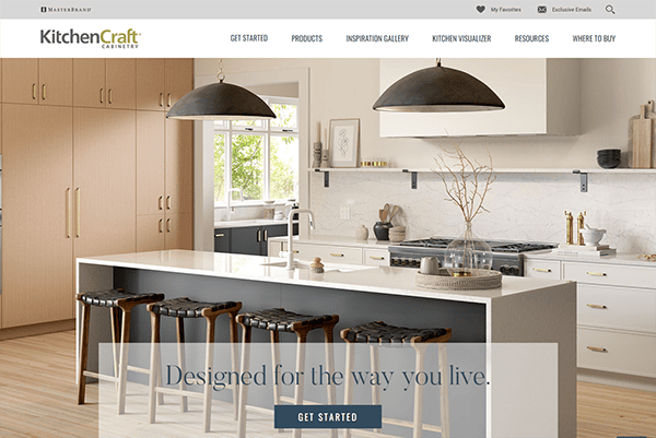 A modern kitchen with an island, black stools, a sink, and a gas stove. Cabinets are light wood, and two large pendant lights hang above the island. The KitchenCraft Cabinetry brand is displayed.