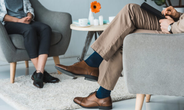 Two individuals sitting in a counseling session. One, likely the psychotherapist, has crossed legs and holds a notepad, while the other is seated with visible shoes. A small table with a flower and cups sits in the background. This cozy setting could be featured on the best websites for mental health resources.