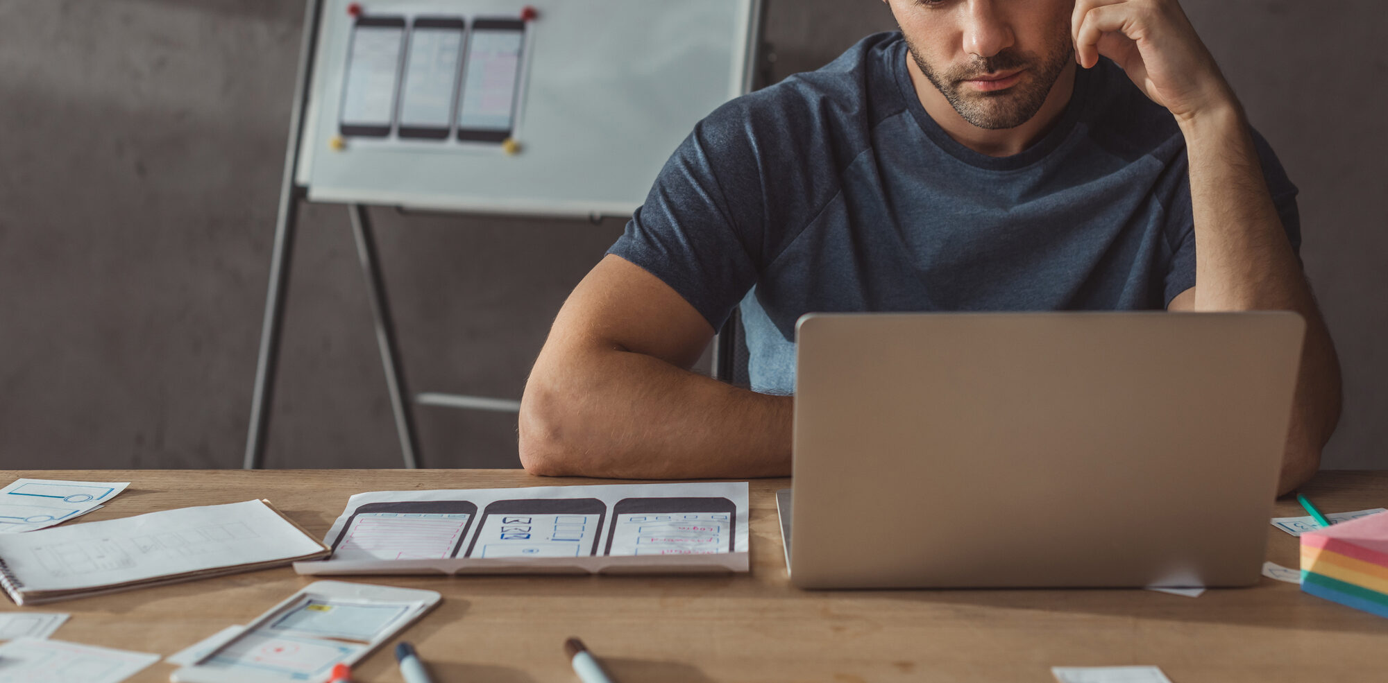 A man sits at a desk working on a laptop, with design sketches and sticky notes spread around, evaluating marketing pricing strategies next to a full whiteboard in the background.