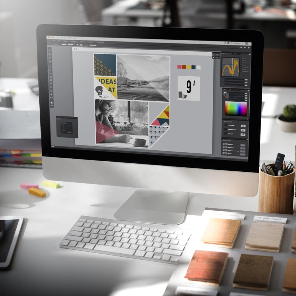 A desktop computer displays graphic design software with a colorful collage, showcasing a Website Redesign in progress. The desk holds a keyboard, mouse, wooden samples, pencils, and office supplies. Other desks are visible in the background.