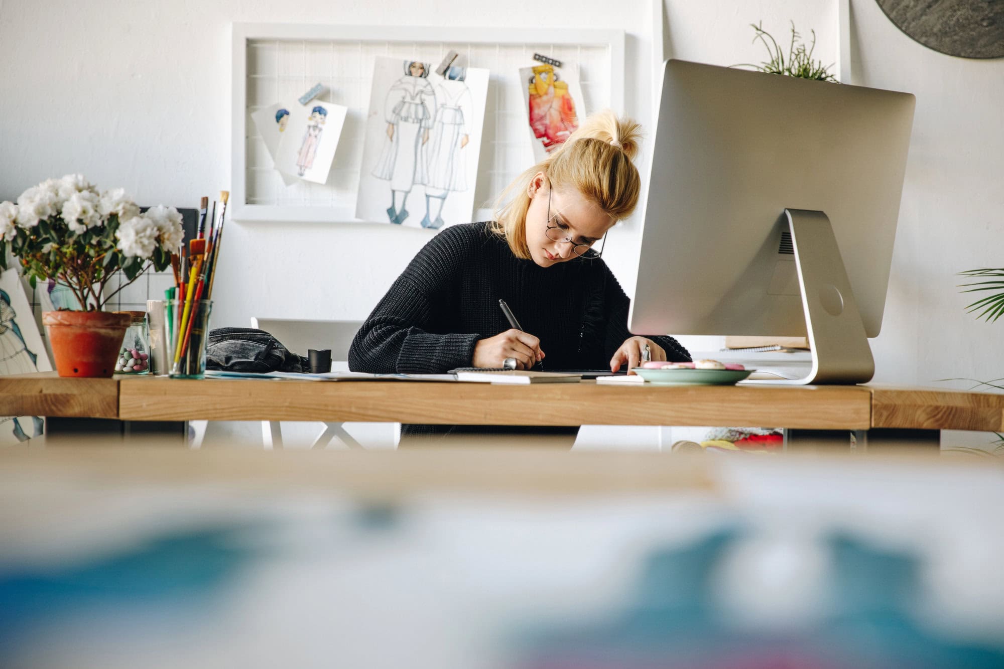 A woman with blonde hair in a black sweater sits at a wooden desk, using a pen tablet alongside a large computer monitor in an art-filled workspace, immersed in providing website redesign services.