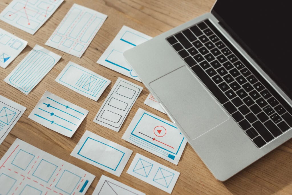 An affordable laptop is placed on a wooden table next to various sketches of web design wireframes and creative layouts.