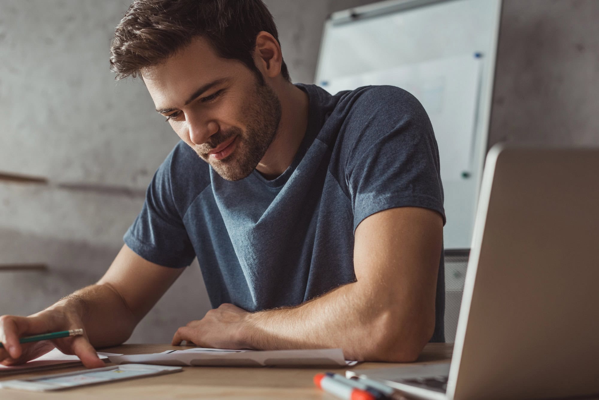A man in a blue shirt sits at a desk, conducting SEO audits on paper while referencing his tablet. A laptop and markers are also on the desk, with a whiteboard in the background.