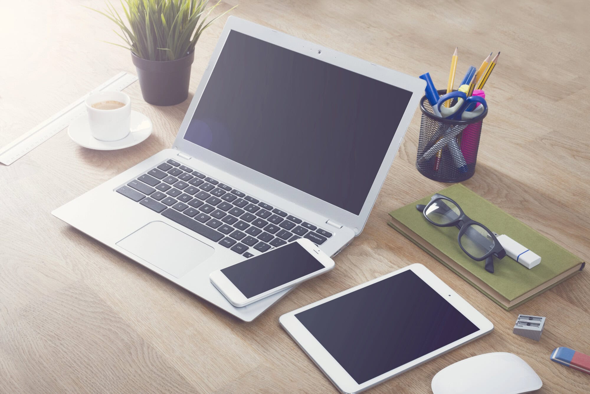 A laptop, smartphone, and tablet on a wooden desk with a cup of coffee, potted plant, and office supplies in the background create the perfect affordable workspace for all your web design needs.