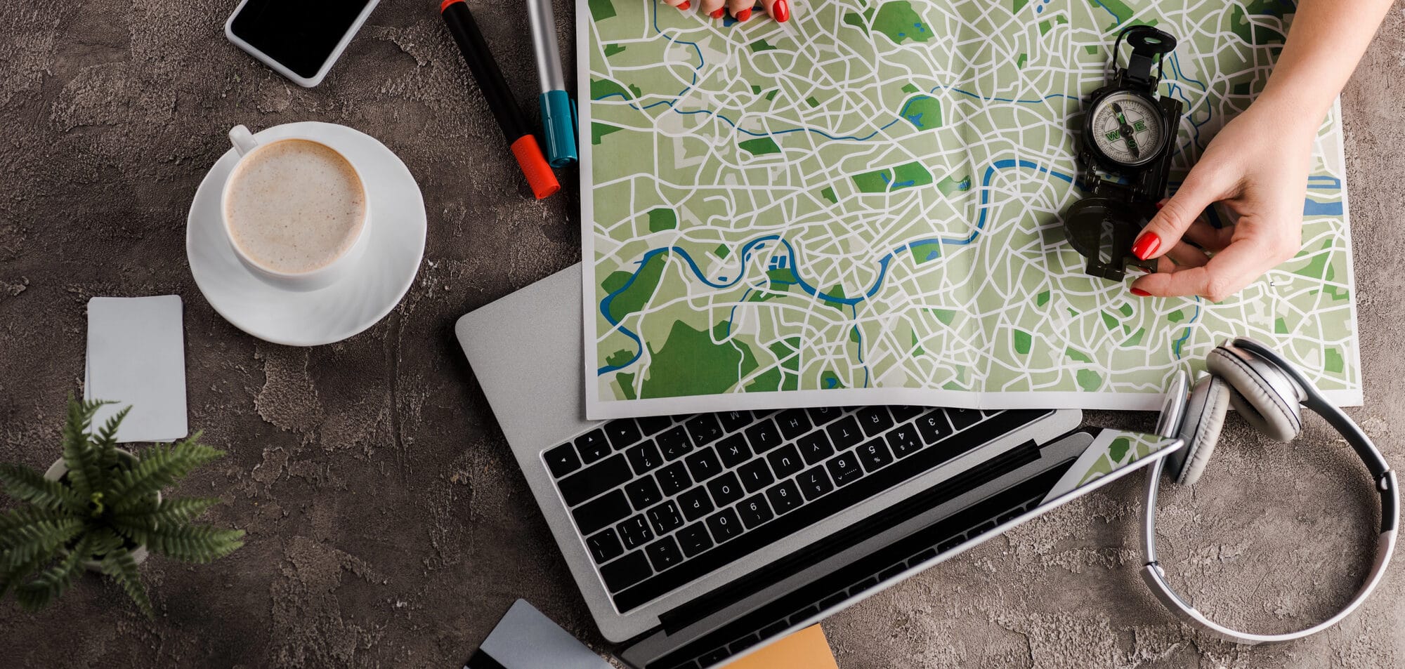 A person holds a map and compass over a laptop, surrounded by a smartphone, headphones, coffee, markers, and note cards on a textured surface—perfect tools for planning an SEO strategy or managing your Google Business profile efficiently.