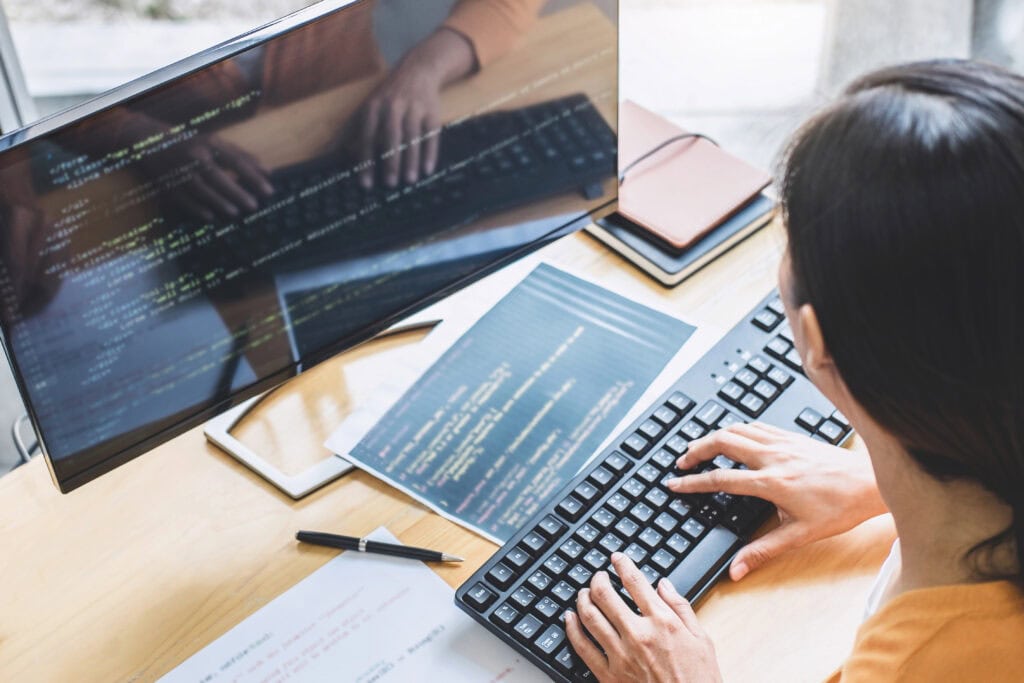A person is typing on a keyboard at a desk, their monitor aglow with programming code and paper documents nearby. Immersed in mastering web design, they navigate both the digital and printed worlds like a pro while consulting their beginner's guide for the core principles of success.
