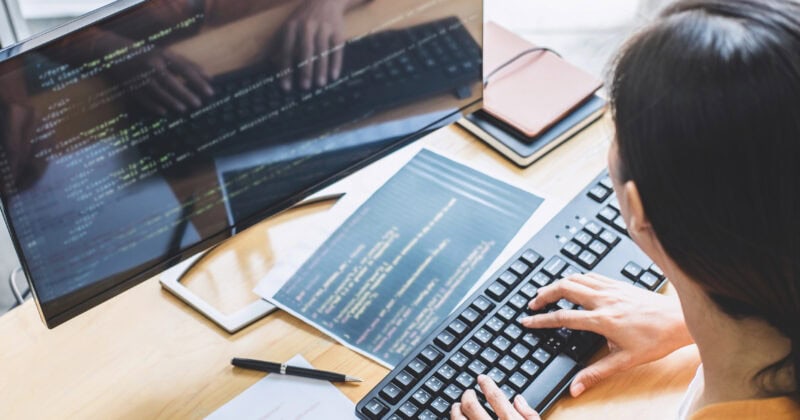 A person is typing on a keyboard at a desk, their monitor aglow with programming code and paper documents nearby. Immersed in mastering web design, they navigate both the digital and printed worlds like a pro while consulting their beginner's guide for the core principles of success.