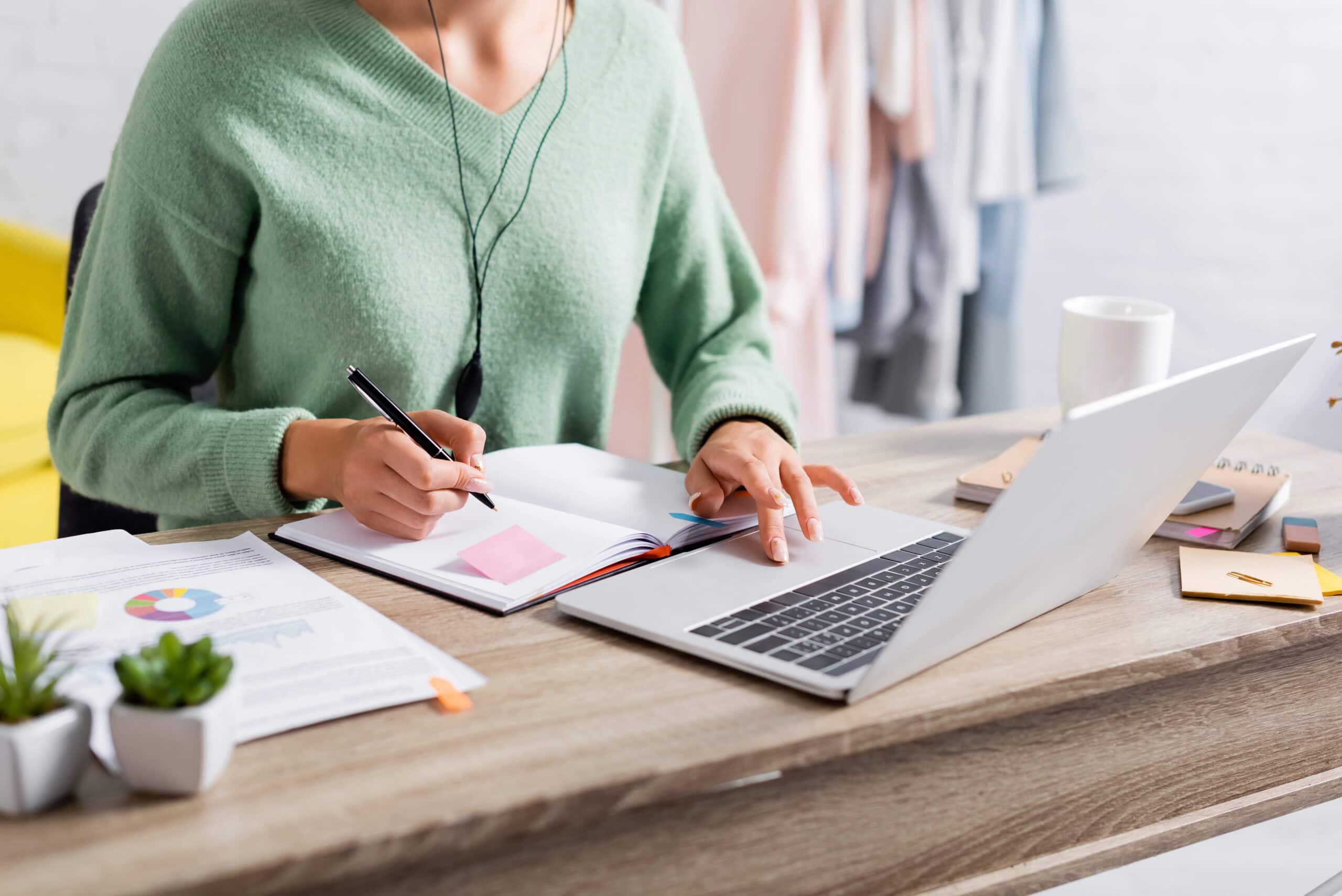A person in a green sweater, focused on marketing strategies, takes notes at a desk while working on a laptop, surrounded by documents and plants.