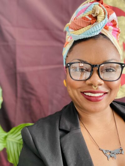 A smiling person wearing glasses and a colorful headwrap, with a septum ring and a necklace shaped like a map. A plant is visible in the background.