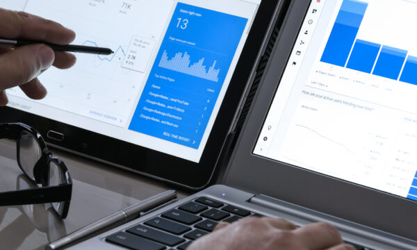 Two people analyze graphs on tablet and laptop screens, one pointing with a pen, optimizing SEO keywords for service businesses. Glasses, smartphone, and pen lie nearby on a table.