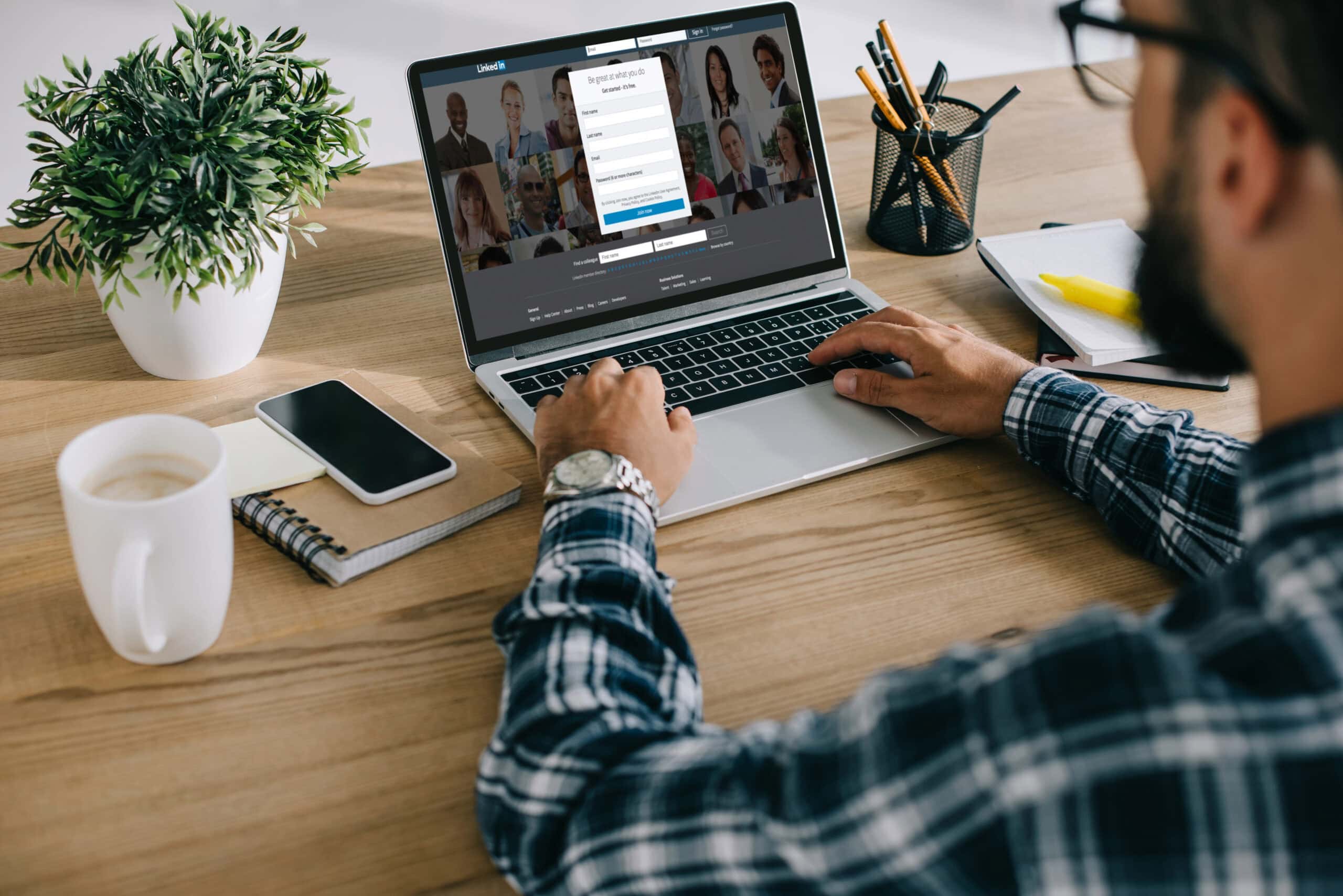 A person wearing a plaid shirt uses a laptop on a wooden desk with a plant, smartphone, notebook, pencils, and glasses. The laptop screen displays high-converting templates for Tree Service Facebook Ads.
