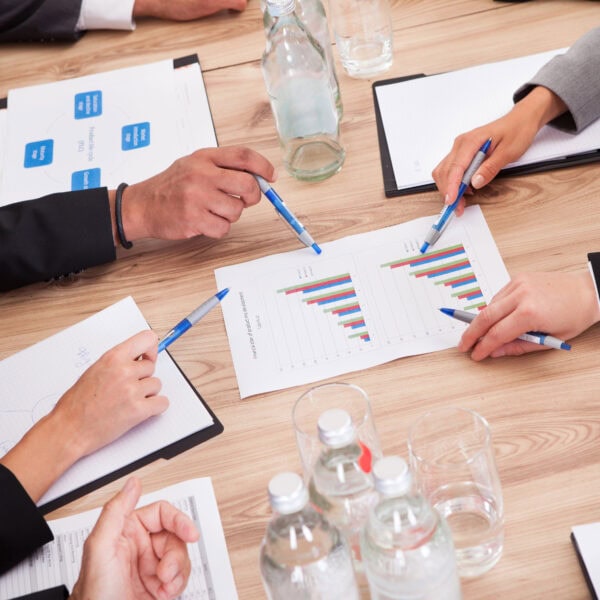 A group of people in business attire review charts and graphs on paper at a table with water bottles, glasses, and notebooks. With pens in hand, they dive into an SEO review, analyzing strategies to boost online visibility.