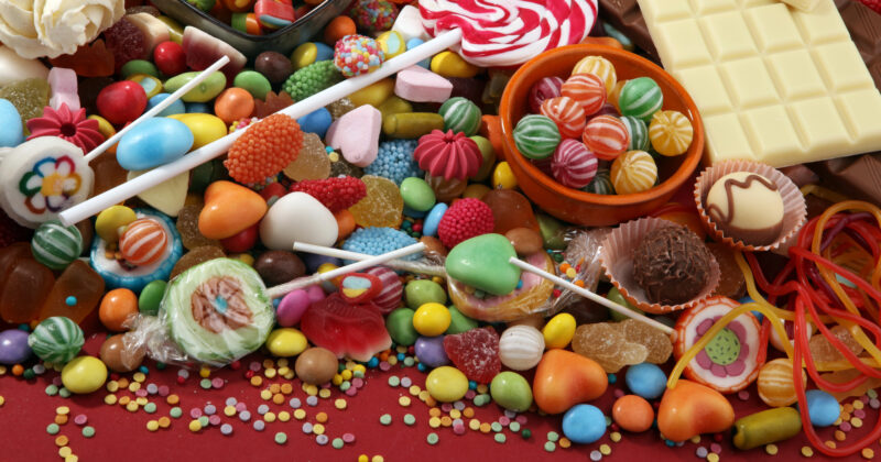 An assortment of the best candy is spread out, including lollipops, chocolate bars, gummies, and colorful sprinkles, on a red surface.