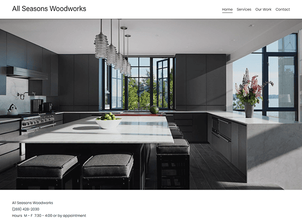 Modern kitchen with large island, bar stools, and pendant lights. Black cabinetry, marble countertops, and large windows overlooking trees. Featured on "All Seasons Woodworks," a leader among the best carpenter websites.