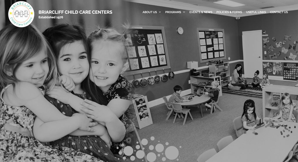 Three young girls smile and hug in the foreground of a classroom at Briarcliff Child Care Center, one of the best daycares, while other children play and interact at small tables and on the floor in the background.