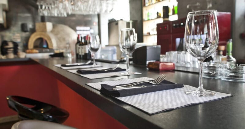 In a stylish restaurant bar setting, empty wine glasses, placemats, and napkins are arranged on a dark countertop. The red and black interior sets the stage for photographers looking to capture scenes that boost marketing strategies, with bottles and glasses artfully displayed in the background.