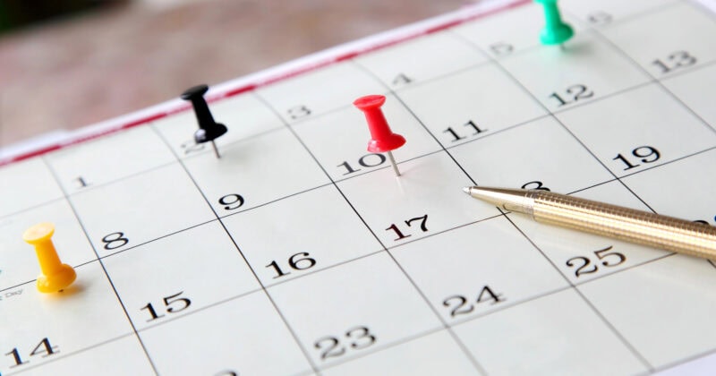 Close-up of a calendar page with dates, perfect for event planners. Red, green, yellow, and black pushpins mark important days. A gold pen rests on the seventeenth.