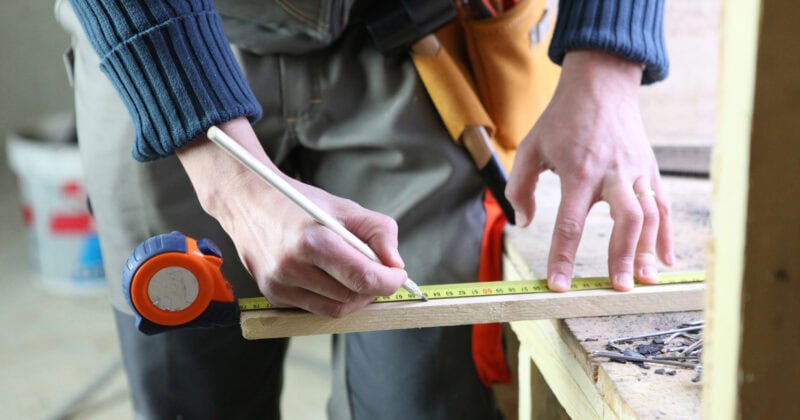 Standing at a workbench filled with tools, a person meticulously measures and marks a wooden plank with precision using a tape measure and pencil. This scene, perfect for a home builder website, highlights the craftsmanship that draws in curious visitors and potential leads.