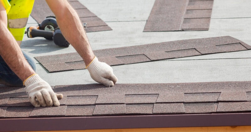 A worker in gloves installs asphalt shingles on a roof, ensuring maximum impact for durability and style.