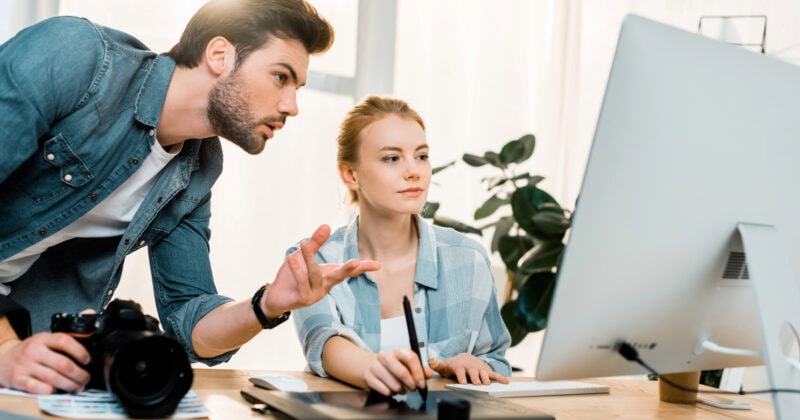 A man and a woman work together at a computer, seamlessly intertwining marketing strategies as the woman uses a graphic tablet. Meanwhile, the man, wielding his camera with the precision of seasoned photographers, focuses intently on increasing exposure to their dynamic project.
