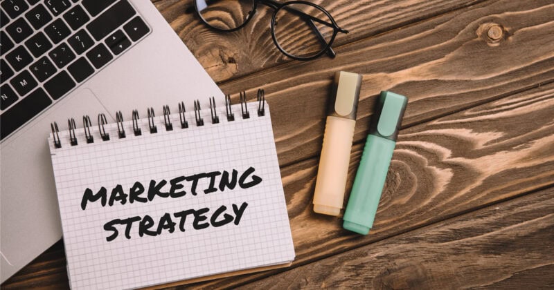 A notebook with "Marketing Strategy" emblazoned on it rests on a wooden table beside a laptop, glasses, and two highlighters, laying the blueprint for building success.