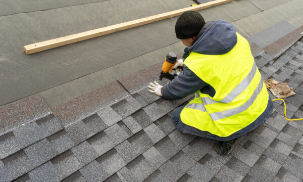 A worker in a yellow safety vest and black beanie uses a nail gun to install asphalt shingles on a roof, surrounded by roofing materials and tools, illustrating the hands-on side of effective roofing marketing.