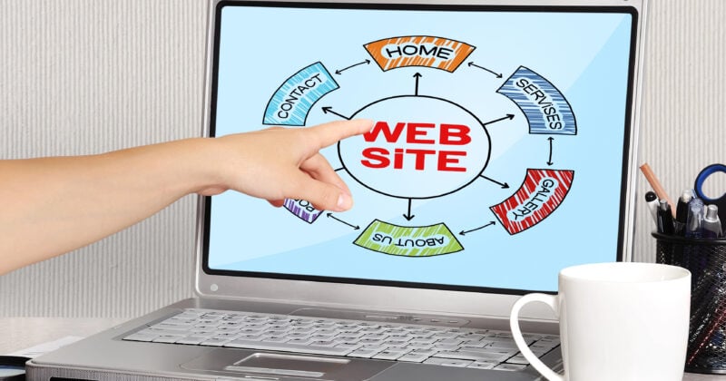 A finger points at a laptop screen showcasing a portfolio-style website structure diagram. Nearby, desk items like a white mug, pens, and a colorful document with charts complete the scene.