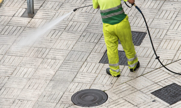 A worker in bright yellow and green reflective clothing uses a high-pressure hose to clean a tiled sidewalk, demonstrating techniques that plumbing Facebook ads use to maximize budget. Only the workers lower body is visible.